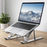 ستاند لاب توب ألومنيوم فضي يدعم جهاز ماك بوك على مكتب خشبي بجانب نبات زينة.
